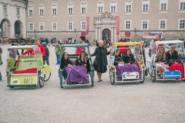 Four differently liveried Rikschatours pedicabs stand side by side on Residenzplatz with passengers under blankets in the seats, the Residenz portal and its Residenzgalerie banners behind.