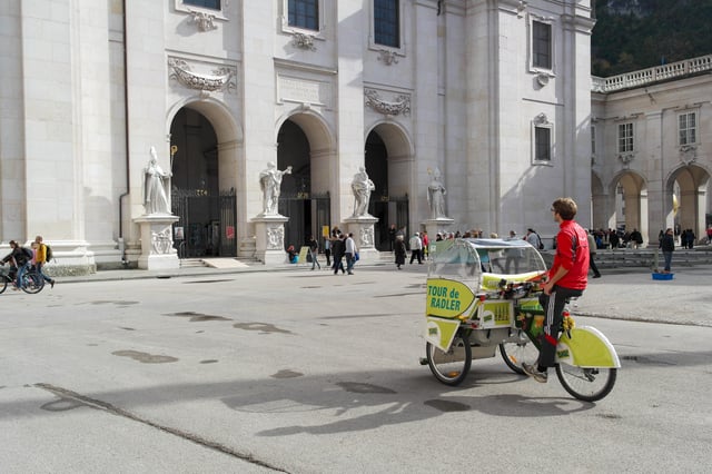 A driver pedals the lime green Sigl Tour de Radler pedicab across Domplatz, the white marble west facade of Salzburg Cathedral with its four saint statues and the dates 774, 1628 and 1959 rising behind.