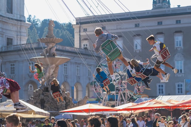 Children and teenagers fly outward on the chain swing carousel at the Rupertikirtag, with the Residenzbrunnen fountain and the Residenz facade behind them and beer garden umbrellas below.
