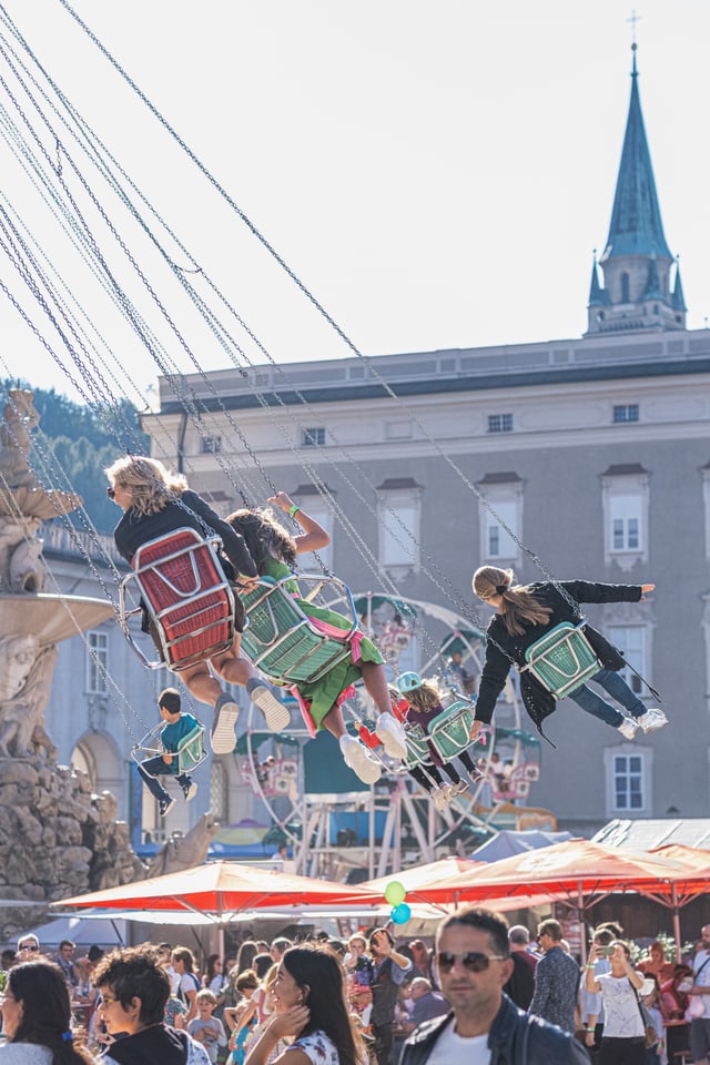 Riders on the chain swing carousel arc out over the crowd while a small painted ferris wheel turns behind them below a slate church spire.
