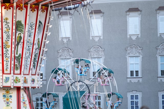 The flower and parrot painted roof of the chain carousel hangs over a small ferris wheel with occupied gondolas, set against the grey stucco facade of the Residenz.