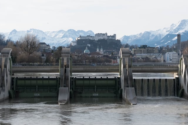 Salzach river weir with Hohensalzburg Fortress and cathedral domes framed by snowy mountains in Salzburg.