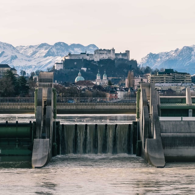 Salzach river weir and waterfall in foreground with Hohensalzburg Fortress and snowy Alps behind.