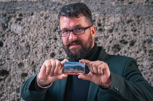 Man in green suit holding a Salzburg Card in front of a stone wall in Salzburg
