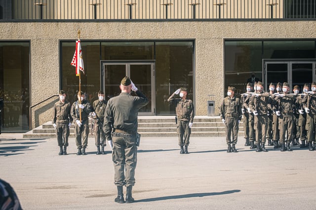 An officer salutes a drawn-up honour guard outside the Festspielhaus entrance, a standard bearer with the red and white Salzburg flag at his left and a rank of soldiers presenting arms at right.