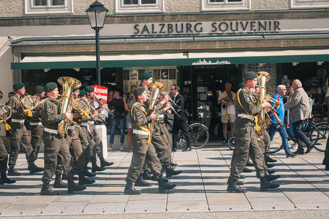 A military band in green berets marches playing tubas, horns and clarinets past the Salzburg Souvenir shop while onlookers watch from the doorway.