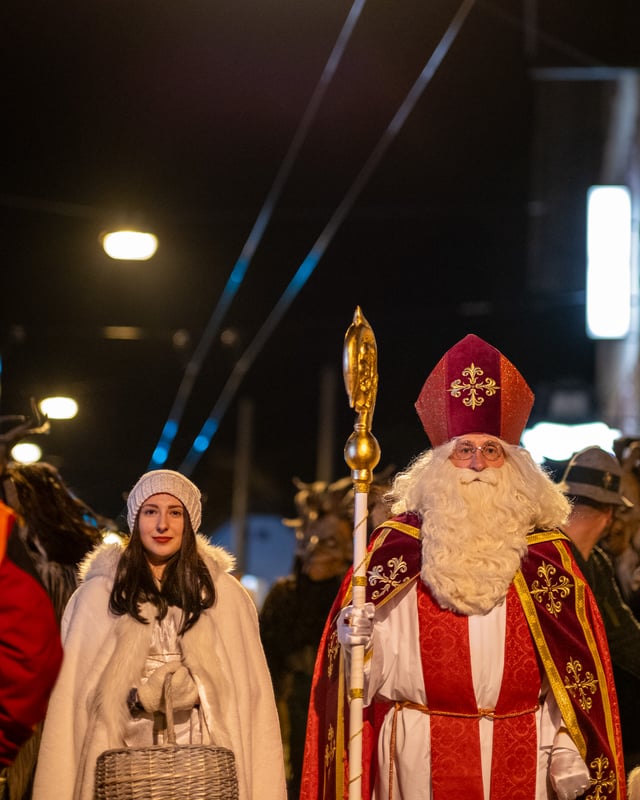 An angel in a white knitted cap and fur trimmed cape holding a wicker basket stands beside Saint Nicholas in mitre, white beard and red and gold cope, with horned masked figures crowding behind them under blue lights.