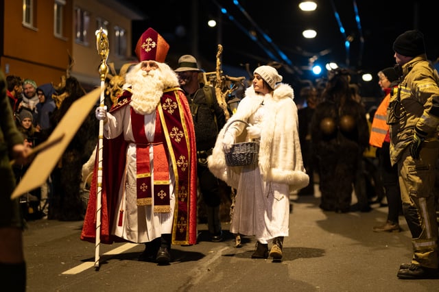 Saint Nicholas in a red and gold cope with a mitre and crozier walks up a village street beside a woman dressed as an angel in a white fur trimmed cape carrying a wicker basket, with masked figures, a lantern bearer and a firefighter behind them.
