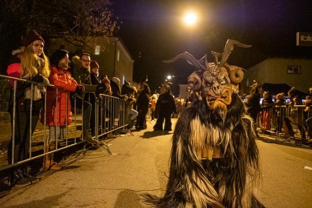 A Krampus with a long black and white mane and tall antler like horns walks down the middle of a village street lit by a single lamp, while spectators in winter coats watch from behind steel barriers on the left.