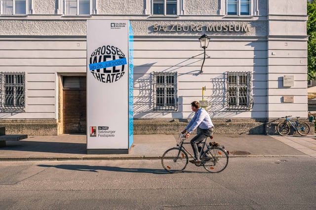 Cyclist passing Salzburg Museum poster column in front of Neue Residenz facade in Salzburg old town