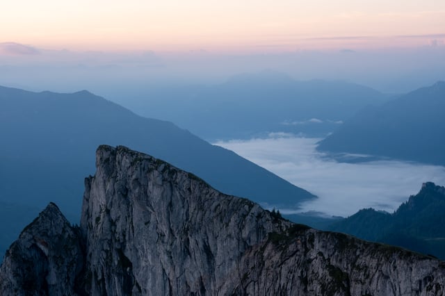 A pale limestone rib of the Schafberg juts into the frame at dawn, with a river of valley fog running below it and blue ridges receding under a soft pink sky.
