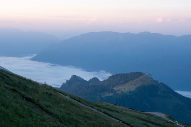 A sea of fog fills the valley below the Schafberg's grassy summit path and its fence, with a lower forested ridge and its scattered alm huts catching the first warm light.
