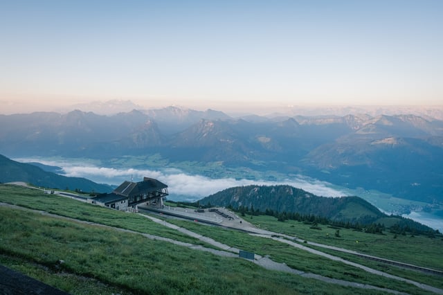 A broad dawn panorama from the Schafberg summit takes in the railway station on its shoulder, the fog-filled Wolfgangsee basin and range upon range of Salzkammergut peaks under an orange horizon.