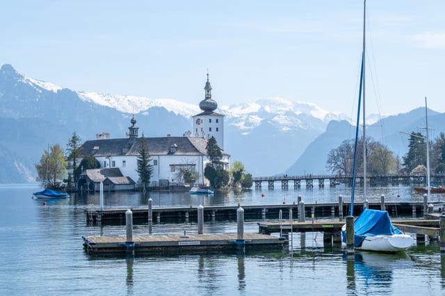 Seeschloss Ort on its island in the Traunsee seen past a wooden boat jetty with tarpaulin-covered sailboats moored in the foreground.