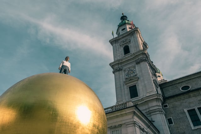 Figure standing on Sphaera sculpture with Salzburg Cathedral clock tower in Salzburg