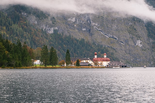 The white pilgrimage chapel of St. Bartholomae with its two red onion domes stands on the shore of the Koenigssee below a huge grey rock face draped in cloud.