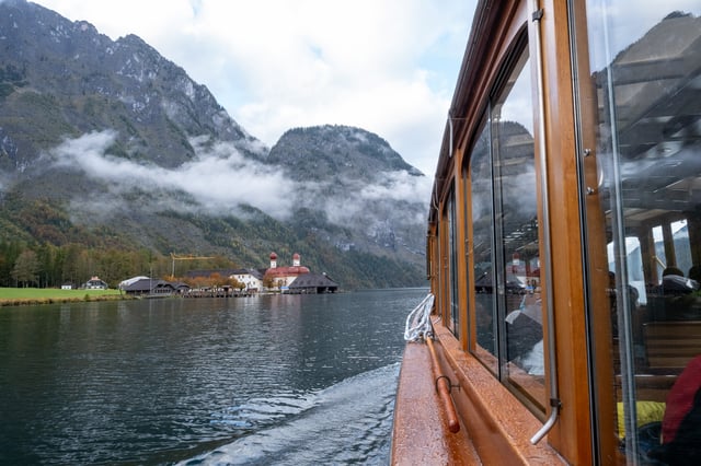St. Bartholomae and its dark boat sheds slide past on the left as the passenger boat moves off, the same scene doubled in the varnished windows on the right.