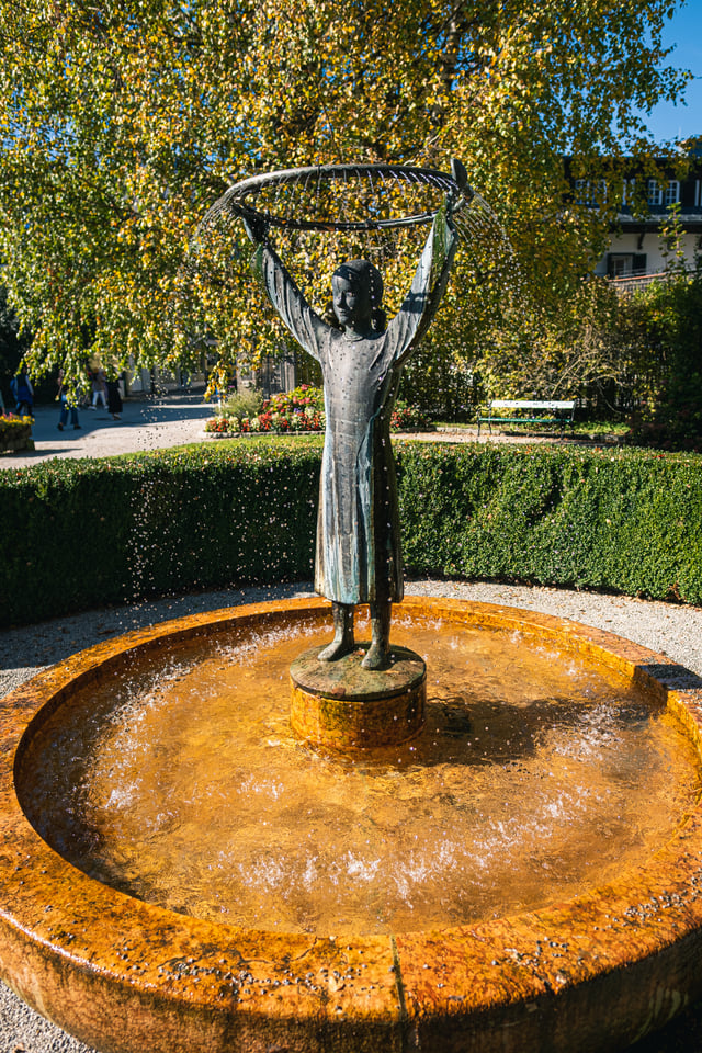 A bronze figure of a girl holds a circular fishing net above her head, water falling from its rim into a round ochre stone basin in a hedged park.