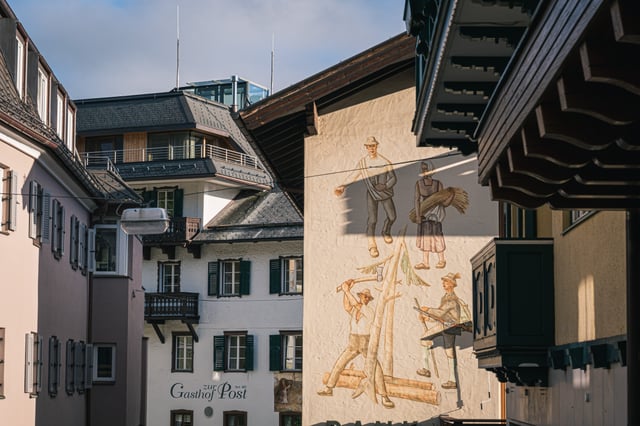 A village gable painted with a sower, a woman carrying sheaves, a woodcutter with an axe and a hunter with a rifle rises beside the white facade lettered Gasthof zur Post seit 1415.