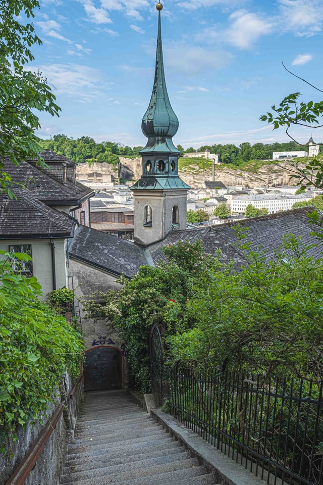 Green onion-dome spire of St John's Church on Imberg seen from stairs descending toward Salzburg old town.