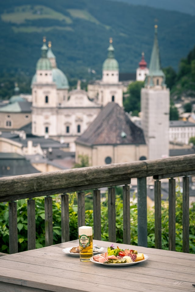 A Brettljause of cold cuts, cheese and pickles with a glass of Wieninger beer on a weathered table at the Stadtalm railing, the domes of Salzburg Cathedral and the Franziskanerkirche tower rising softly behind.