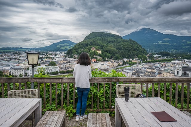 A woman stands with her back to the camera at the wooden railing of the Stadtalm terrace on the Mönchsberg, looking across the Salzburg Neustadt to the Kapuzinerberg with its monastery and the cloud-capped Gaisberg beyond.