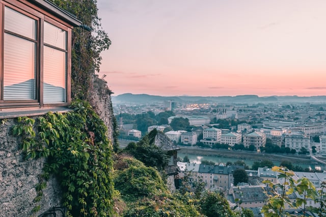 Pink sky over Salzburg seen from the Stadtalm on the Mönchsberg, with the ivy-covered rock wall and a shingled turret in the foreground and the Salzach curving past the Neustadt rooftops below.