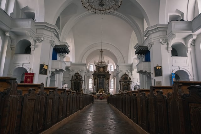 The central aisle of Hallein parish church runs between dark wooden pews toward the altar, under a white barrel vault hung with two crystal chandeliers.