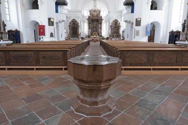 An octagonal red marble baptismal font with a hammered metal lid stands on the tiled floor of Hallein parish church, the pews and altar receding behind it.