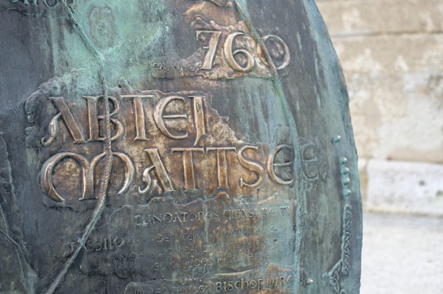 Close-up of a green-patinated bronze bell cast in raised lettering with the words Abtei Mattsee and the year 760 above a Latin and German inscription naming the founders, standing against a pale stone wall.