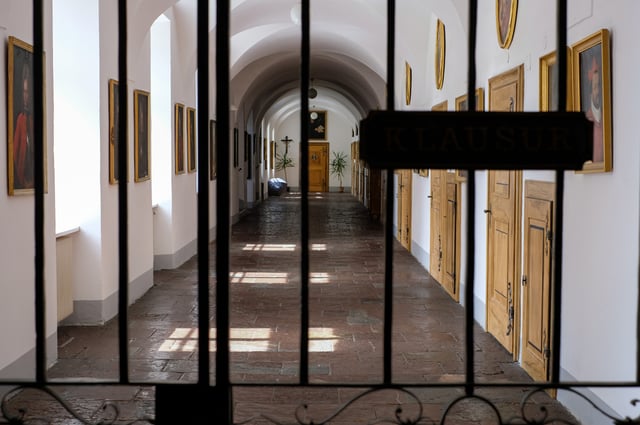 An iron grille carrying a painted KLAUSUR sign closes off a sunlit corridor of pale timber doors and dark portrait paintings inside the Mattsee collegiate foundation.