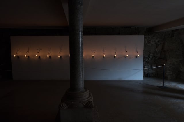 A wide dark view of the crypt beneath Salzburg Cathedral with a row of candle brackets and skeleton figures glowing along two pale panels behind a Romanesque column.