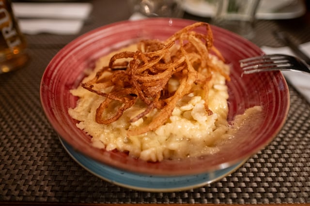 A red rustic plate of cheese Spätzle crowned with a nest of crisp fried onions, shot close and low with a beer glass out of focus behind it on a woven placemat.