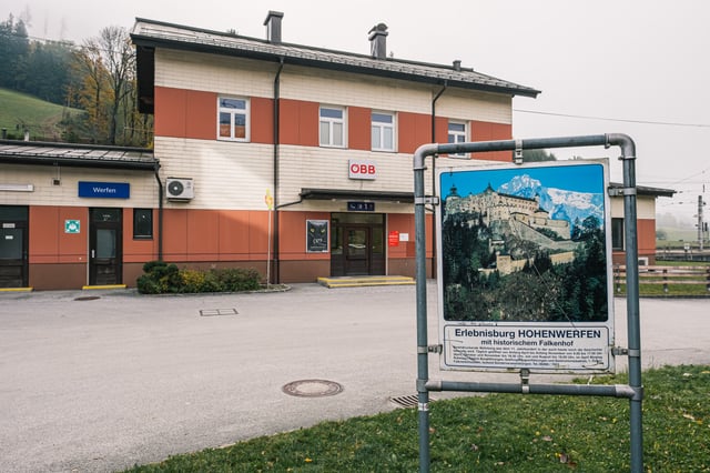 The cream and red Werfen station building seen from the forecourt, with the OeBB logo above the entrance and a weathered Hohenwerfen fortress billboard standing on the grass to the right.
