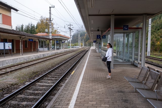 A woman in a white jacket waits alone on platform 2 at Werfen station on a misty autumn morning, with empty tracks running away between wooded slopes.