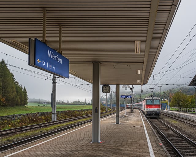 Under the platform canopy at Werfen station a blue Werfen sign hangs beside the clock while a red and white OeBB locomotive draws its train in along the far track on a misty morning.