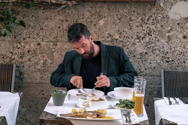 A bearded man in a dark jacket eats Tafelspitz at a white-clothed terrace table set against the pebbly conglomerate wall of the Hotel Elefant building, with a trout, a salad and a wheat beer on the table.