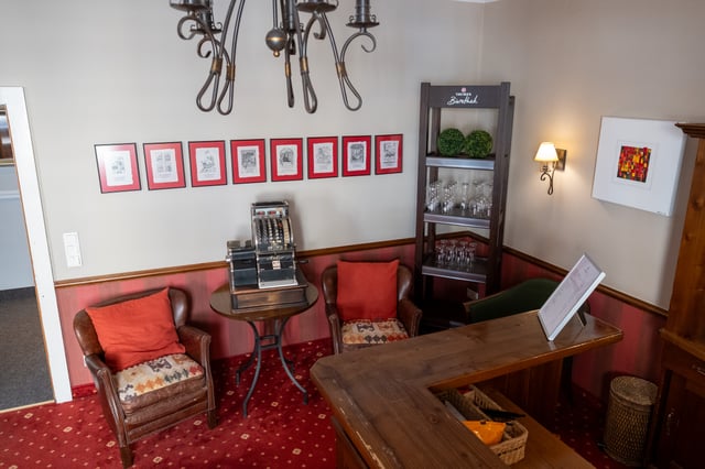 The reception corner of a Salzburg hotel bar, with an antique Anker cash register on a bistro table between two leather armchairs, a row of red-mounted Salzburg prints and a Trumer Bierothek glass shelf.