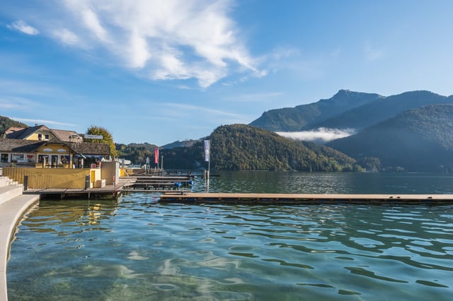 Wolfgangsee lies glassy in low morning sun, seen past the St. Gilgen landing stage, with a band of cloud caught on a wooded ridge below a rocky summit.