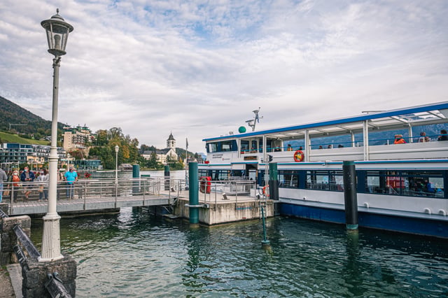 A Wolfgangsee Schifffahrt passenger ferry lies at the St. Wolfgang landing stage while masked passengers wait on the jetty, the village church tower rising across the water.