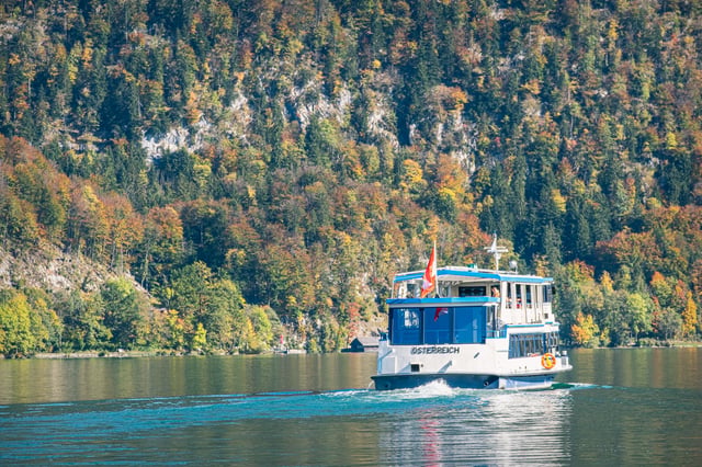The ferry MS Österreich runs along the steep autumn-coloured shore of the Wolfgangsee, leaving a wake across the green water.