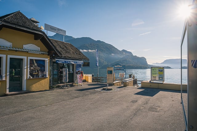 The yellow ticket house at the St Gilgen pier, signed Wolfgangsee Schifffahrt and Schafbergbahn above a Kassa window with a postcard stand outside, stands empty in low morning light with the lake and misted ridges beyond.