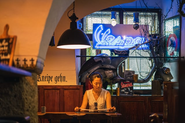 A guest reads the menu under an industrial pendant lamp at Zwettler's Wirtshaus in Salzburg, with a mounted Vespa scooter and its blue neon sign filling the wall behind her.