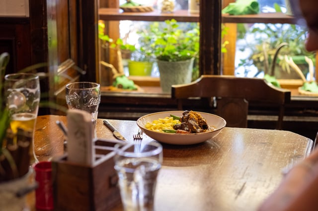 A bowl of beef goulash with ribbon noodles sits on a wooden table by a window full of potted plants at Zwettler's Wirtshaus in Salzburg.