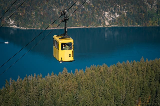 A yellow cabin numbered 3 of the old Zwölferhorn cable car crosses high above the deep blue Wolfgangsee while a passenger leans out of the window with a camera.
