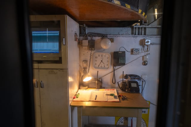 The cable car operator's corner desk at the Zwölferhorn station holds a lamp-lit logbook, handheld radios and telephones, with a wall clock and an old monitor listing ride records above it.